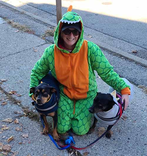 belmont staff member wearing dinosaur costume kneels on ground with two dogs